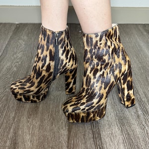 May include: A pair of platform ankle boots with a leopard print pattern. The boots have a zipper closure and a chunky heel. The platform sole and heel add height. The boots are made of a textured material.