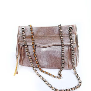 May include: A metallic rose gold handbag with a chain strap. The bag has a textured finish and a flap closure. The chain strap is silver with a brown leather detail. The bag has a rectangular shape and a small tassel detail.