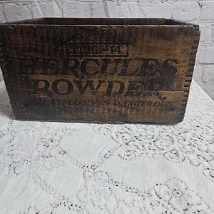 Könnte beinhalten: Eine rechteckige, dunkelbraune Holzkiste mit gezinkten Ecken. Die Vorderseite zeigt schwarzen Schablonentext mit der Aufschrift HERCULES POWDER und EXPLOSIVES DANGEROUS sowie ein kleines Feld mit dem Text LC-14 über der Hauptbeschriftung.