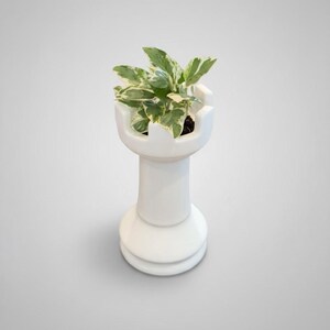 May include: A white ceramic chess rook planter holding a small green and white plant. The planter has a glossy finish and is set against a light gray background. The plant's leaves have a variegated pattern.