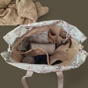 May include: Open tote bag with a floral pattern in light blue and pink. The bag is filled with yarn, knitting needles, and other craft supplies. A beige knitted shawl is visible in the background. The bag has beige handles.