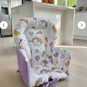 Può includere: Seggiolino per bambini con cuscino bianco con motivo unicorno e arcobaleno. Il seggiolino ha un bordo trapuntato lilla e un fiocco di nastro lilla. Progettato per comfort e supporto.
