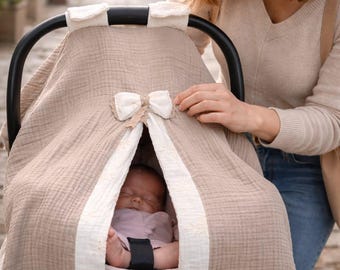 May include: A beige baby car seat cover with a white bow and trim. The cover is draped over the car seat, with a sleeping baby visible inside. The cover has a ruffled edge and is being held by a person.