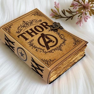 Puede incluir: Un diario de cuero marrón con la palabra "THOR" y el logotipo de los Vengadores. El diario tiene un borde decorativo y un cierre de metal. Las páginas tienen un aspecto envejecido, dándole un aspecto vintage.