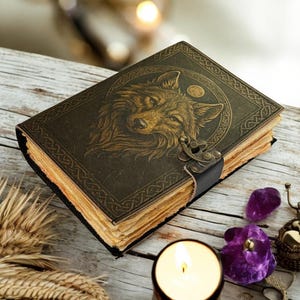 May include: A leather-bound journal with a wolf design on the cover. The cover is dark brown with an embossed wolf head and circular design. The pages have a vintage, aged look. A black leather strap and metal clasp secure the journal.
