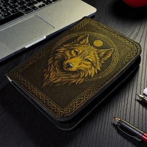 Puede incluir: Cartera de cuero marrón oscuro con una ilustración dorada de un lobo en la parte delantera. El lobo está rodeado por un diseño circular con una luna. La cartera tiene un borde decorativo.