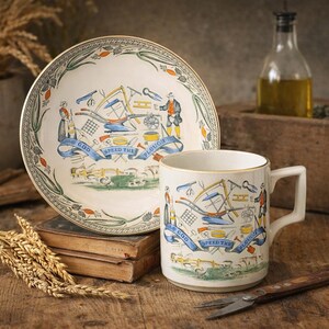 May include: A vintage-style ceramic plate and mug set. The plate and mug feature a detailed illustration with farming tools, figures, and the text "GOD SPEED THE PLOUGH" on a blue banner. The set has a cream background with a gold rim.