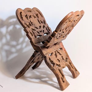 May include: A wooden book stand with intricate, openwork carvings. The stand is made of a warm, medium-toned wood and has a folding design. The carved details include floral and leaf patterns, adding a decorative element to the functional piece.