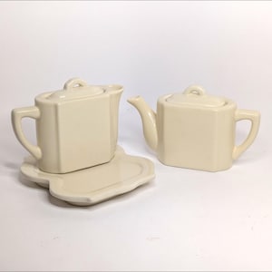 May include: Two off-white ceramic teapots with matching lids and handles. One teapot sits on a matching saucer. The teapots have a geometric design and are suitable for serving tea.