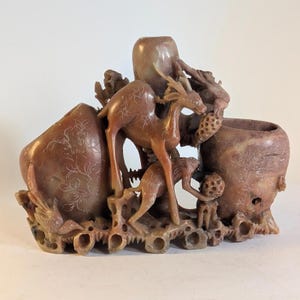May include: A detailed, reddish-brown carved stone sculpture featuring deer and other animals. The sculpture has a complex design with multiple compartments and intricate floral patterns. The overall theme is nature and wildlife.