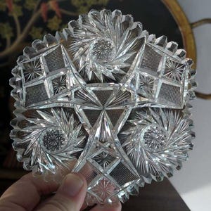 May include: A clear, circular glass dish featuring an intricate, multi-faceted cut pattern. The design includes three prominent starburst motifs alternating with geometric sections of cross-hatched grid lines and triangular prisms.