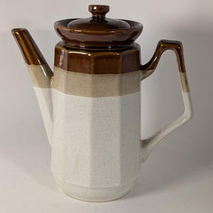 May include: A vintage ceramic coffee pot with a brown lid and handle, a brown and tan upper section, and a white speckled lower section. The pot has a geometric design and a spout for pouring.