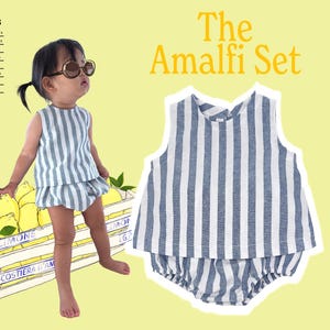 Op de afbeelding: De Amalfi Set, een baby-outfit met blauw-witte strepen, wordt getoond. De set bestaat uit een mouwloze top en bijpassende shorts. Een kind draagt de outfit, naast een kist met citroenen. De tekst bevat maten en "The Amalfi Set".