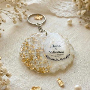 Puede incluir: Un llavero de resina blanca y dorada con los nombres "Anna & Sebastian" y la fecha "18 de septiembre de 2023" impresos. El llavero tiene un llavero y una cadena plateados. Acentos de pan de oro en la resina. El fondo incluye perlas y pan de oro.