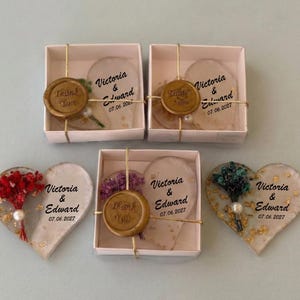 以下が含まれることがあります： ハート型の結婚式の記念品。ドライフラワーと金色のアクセントが特徴です。「Victoria & Edward」と日付「07.06.2027」でパーソナライズされています。一部は白い小箱に梱包されています。