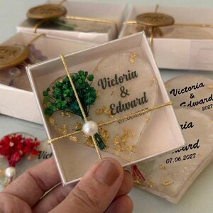 以下が含まれることがあります： 「Victoria & Edward」の名前と「07.06.2027」の日付が印刷されたハート型の結婚式の記念品。各記念品は金箔と小さな緑色の花のブーケで飾られています。記念品は白い箱に梱包されています。