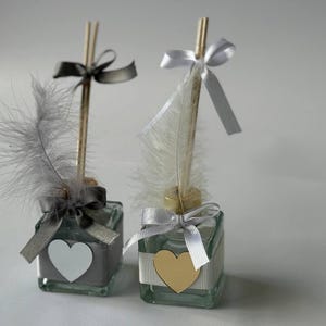 May include: Two decorative glass bottles with reed diffusers. Each bottle has a ribbon-tied bow, a feather, and a heart-shaped accent. One heart is white, the other is gold. The bottles are filled with a gray liquid.