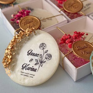 以下が含まれることがあります： アイボリー色の結婚式の引き出物。金箔のアクセントと花のディテールが特徴です。「Issac & Karina」の名前と「DECEMBER 7, 2024」の日付が記載されたパーソナライズされたメッセージが各引き出物にあります。「Thank You」のシールが付いた白い箱に入っています。
