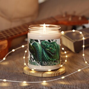 May include: A clear glass jar candle with a white wax fill and a lit wick. The label features a detailed illustration of a green snake coiled on a branch with the text "SNAKE MODE ON" printed in a bold, light-colored font at the bottom.