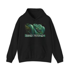 May include: A black hooded sweatshirt featuring a graphic print of a coiled green snake on a tree branch. The text SNAKE MODE ON is printed in white capital letters below the snake illustration. The hoodie includes a front kangaroo pocket.