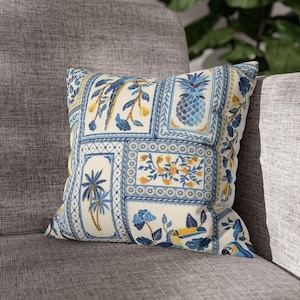 Può includere: Un cuscino quadrato con sfondo color crema e un motivo a piastrelle blu e giallo. Il design include illustrazioni di ananas, palme, uccelli tropicali e motivi floreali disposti all'interno di bordi rettangolari.