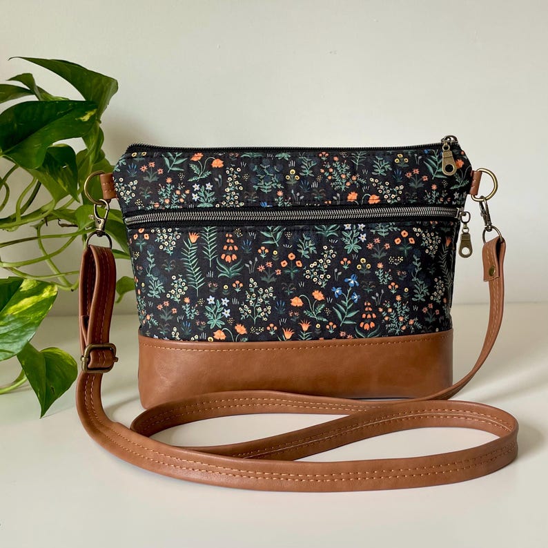 May include: A crossbody bag with a floral pattern on a black background, featuring orange, green, and blue flowers. The bottom is brown leather, and it has a brown leather strap and silver zippers.