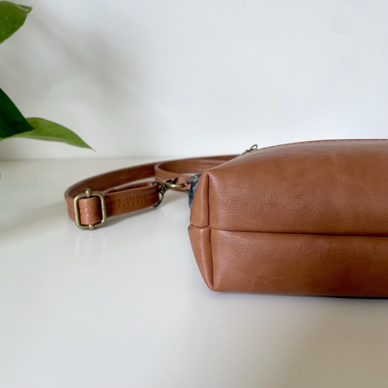 May include: A brown leather crossbody bag with a matching adjustable strap and a brass-colored buckle. The bag has a rectangular shape with rounded corners and a zipper closure. The bag is sitting on a white surface.