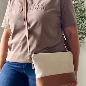 May include: A brown t-shirt with the word "MAMA" embroidered on the front. A crossbody bag with a beige upper and brown bottom is worn. The bag has a long, thin strap.