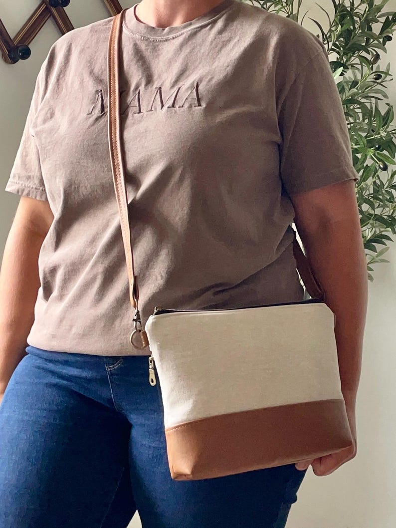 May include: A brown t-shirt with the word "MAMA" embroidered on the front. A crossbody bag with a beige upper and brown bottom is worn. The bag has a long, thin strap.