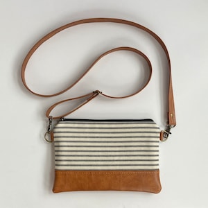 May include: A crossbody bag with a striped fabric body and a brown leather bottom. The bag has a long, adjustable brown leather strap and a black zipper closure. The hardware is a brass color.