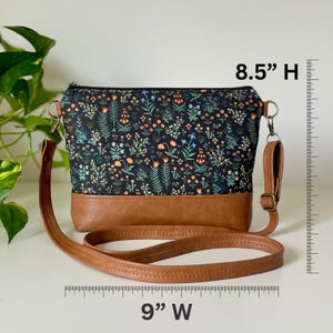 May include: A crossbody bag with a floral pattern on a black background and a brown leather base. The bag is 9 inches wide and 8.5 inches high, with a brown leather strap and gold-colored hardware.