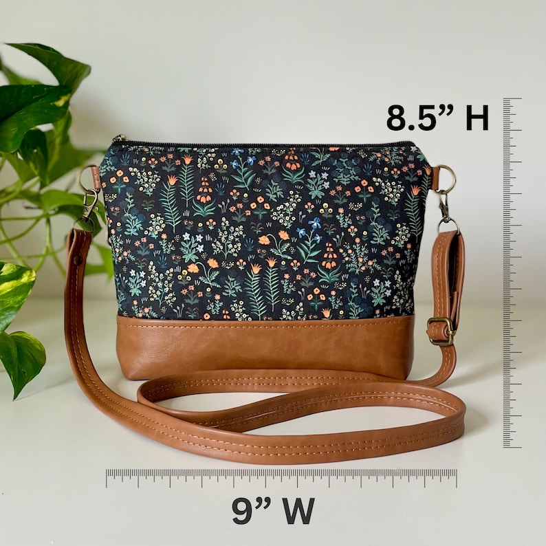 May include: A crossbody bag with a floral pattern on a black background and a brown leather base. The bag is 9 inches wide and 8.5 inches high, with a brown leather strap and gold-colored hardware.