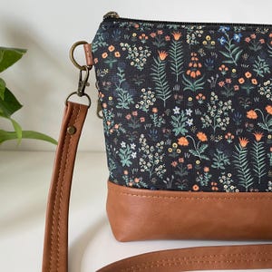 May include: A floral print crossbody bag with a brown leather base and strap. The bag features a black background with a colorful floral pattern. The strap is attached with a metal clasp. The bag is approximately 9 inches wide.