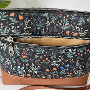 May include: A black floral print crossbody bag with a brown leather bottom. The bag features a zipper closure and a tan interior. The floral pattern includes orange, blue, and white flowers and green leaves.
