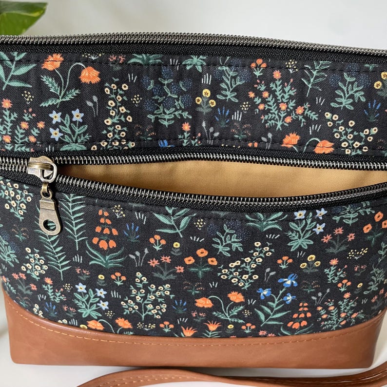 May include: A black floral print crossbody bag with a brown leather bottom. The bag features a zipper closure and a tan interior. The floral pattern includes orange, blue, and white flowers and green leaves.