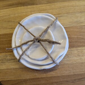 May include: A stack of three small, off-white ceramic plates tied with twine. The plates have an irregular, organic shape. The twine is wrapped around the plates, holding them together. The plates are on a wooden surface.