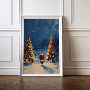May include: Framed Christmas art print. The painting shows Santa Claus walking on a snowy path between two illuminated Christmas trees. The background is a dark blue night sky with falling snow. The frame is white.