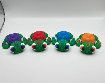Peut inclure: Quatre figurines de tortues colorées. Chaque tortue a un corps vert et une carapace de couleur différente : violet, rouge, bleu et orange. Les tortues ont des yeux blancs et noirs et sont disposées en rangée.