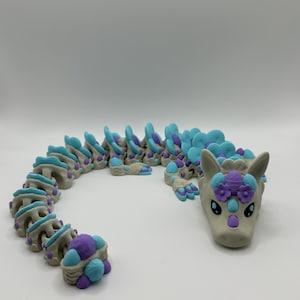 May include: A flexible, articulated dragon figurine in light gray, teal, and purple. The dragon's head features a floral crown. The body is segmented, allowing for movement. The dragon's design is whimsical and colorful.