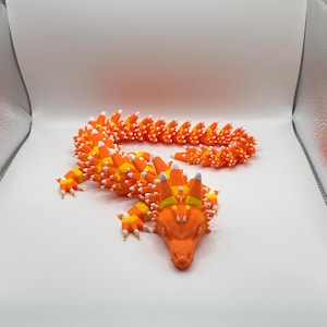May include: A 3D-printed orange dragon figurine with a candy corn design. The dragon's body is segmented, with orange, yellow, and white details. The head is orange with yellow horns and white accents. The dragon is posed on a white surface.