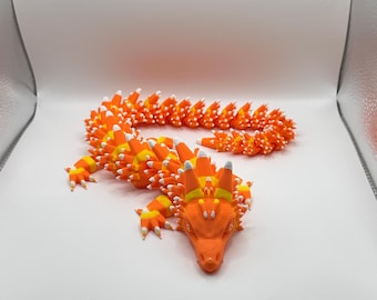 May include: A 3D-printed orange dragon figurine with a candy corn design. The dragon's body is segmented, with orange, yellow, and white details. The head is orange with yellow horns and white accents. The dragon is posed on a white surface.