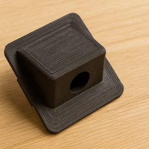 Può includere: Un componente quadrato nero opaco stampato in 3D, caratterizzato da un blocco centrale rialzato con un foro circolare su un lato. L'oggetto presenta una distinta texture a strati sulla superficie, che indica il processo di produzione additiva.