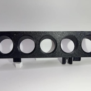 Peut inclure: Un panneau de jauge rectangulaire noir comportant cinq découpes circulaires disposées sur une rangée horizontale. La surface présente une finition mate et texturée. Deux petits supports de montage dépassent du bord inférieur du panneau.