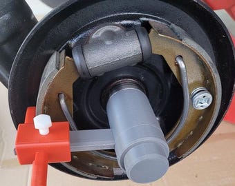 May include: Close-up of a car's brake assembly, featuring a black drum, metallic components, and a gray cylinder. A red and white tool is attached, likely for maintenance or adjustment. The image highlights automotive parts and repair.