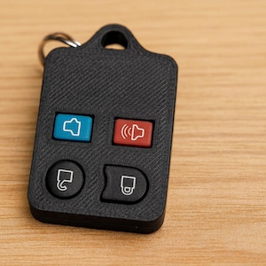 May include: Black car key fob with four buttons. The buttons are colored blue, red, and black. The key fob has a key ring attached to the top. The background is a light brown wooden surface.