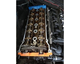 May include: An overhead view of an engine's cylinder head, showcasing the camshafts, valves, and timing chain. The components are metallic with a mix of gold and silver tones, set against a dark background. Orange and blue accents are visible.