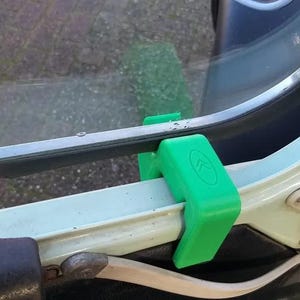 Peut inclure: Un clip en plastique vert vif fixé sur le cadre de la fenêtre d'une portière de véhicule de couleur claire. Le clip présente un petit logo en relief composé de deux chevrons pointant vers le haut sur la surface supérieure de la pièce.