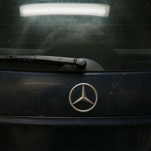 Può includere: Primo piano del bagagliaio di un'auto blu scuro con un emblema Mercedes-Benz argentato. Il lunotto ha un tergicristallo ed è leggermente sporco. L'immagine è ben illuminata.