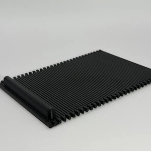 Könnte beinhalten: Eine rechteckige schwarze Kunststoffmatte mit einer Reihe paralleler, dünner, erhabener Rippen auf der Oberfläche. Eine Seite der Matte enthält einen dickeren, massiven zylindrischen Stab, der über die gesamte Breite verläuft.