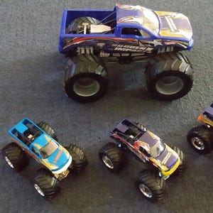 K&ouml;nnte beinhalten: Eine Sammlung von Spielzeug-Monstertrucks in verschiedenen Farben, darunter Blau, Gelb und Lila. Die Trucks haben gro&szlig;e, schwarze Reifen und detaillierte Designs. Ein Truck ist gr&ouml;&szlig;er als die anderen.